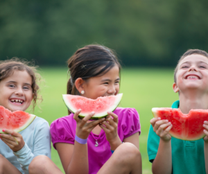 Three young children sitting outside on grass, laughing and smiling while eating large slices of watermelon. The background is green and slightly blurred, suggesting a warm, sunny day.