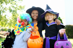 Three children are standing outside. A child dressed up as a clown and two children dressed up as witches. The witches are holding pumpkin candy buckets. 