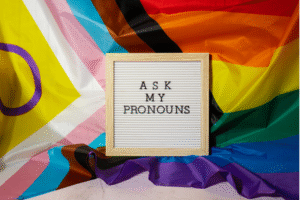 A sign stating "Ask my pronouns" in front of the activist Pride flag.