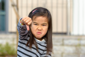 A young child with long dark hair wearing a striped shirt is making an angry face and giving a thumbs-down gesture.