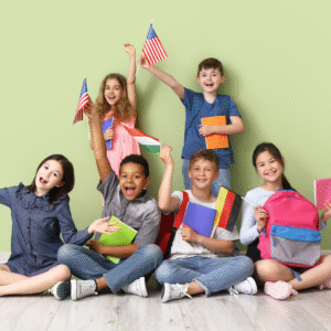 A group of six smiling children sit together on the floor in front of a light green wall, holding colorful notebooks, backpacks, and small flags from different countries, including the United States, Hungary, and Germany. The children appear cheerful and excited, celebrating diversity and learning.