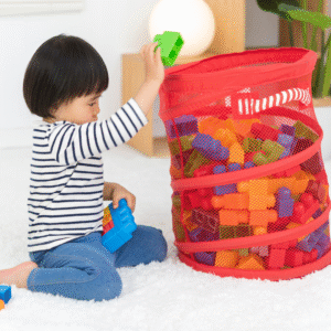 A young child wearing a striped long-sleeve shirt and blue pants sits on a white carpet, placing a green toy block into a large red mesh basket filled with colorful plastic building blocks. The scene appears calm and focused, showing the child cleaning up toys during playtime.