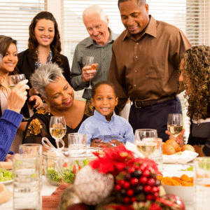 A multigenerational family gathers around a holiday table, smiling and talking together. An older woman has her arms around a young boy who is smiling at the camera. Other adults stand and sit nearby holding glasses, with festive food and decorations on the table in the foreground.