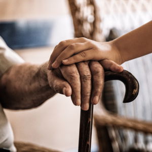 Close-up of a younger person's hand gently resting on top of an older person's hand as they hold a wooden cane, conveying comfort and support.