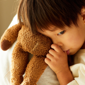 A young child rests their face against a worn brown stuffed animal while gently sucking their thumb. The child looks downward with a quiet, sad expression, holding the toy close for comfort.