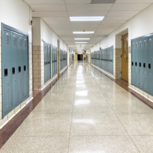 A long, empty school hallway with rows of blue lockers lining both sides, fluorescent ceiling lights overhead, and a shiny tile floor reflecting the lights, creating a quiet, symmetrical scene.