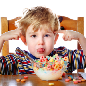 Young child sitting at a table with a bowl of colorful cereal and milk, sticking out their tongue and pointing to their temples with both hands, cereal pieces scattered on the table.