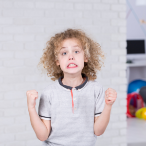 A young child with curly hair clenches their fists and makes a tense, frustrated facial expression, as if feeling angry or overwhelmed, while standing indoors against a light brick wall background.