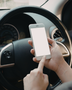 Person holding a smartphone with a blank screen while sitting behind the steering wheel of a car, with dashboard and controls visible in the background.